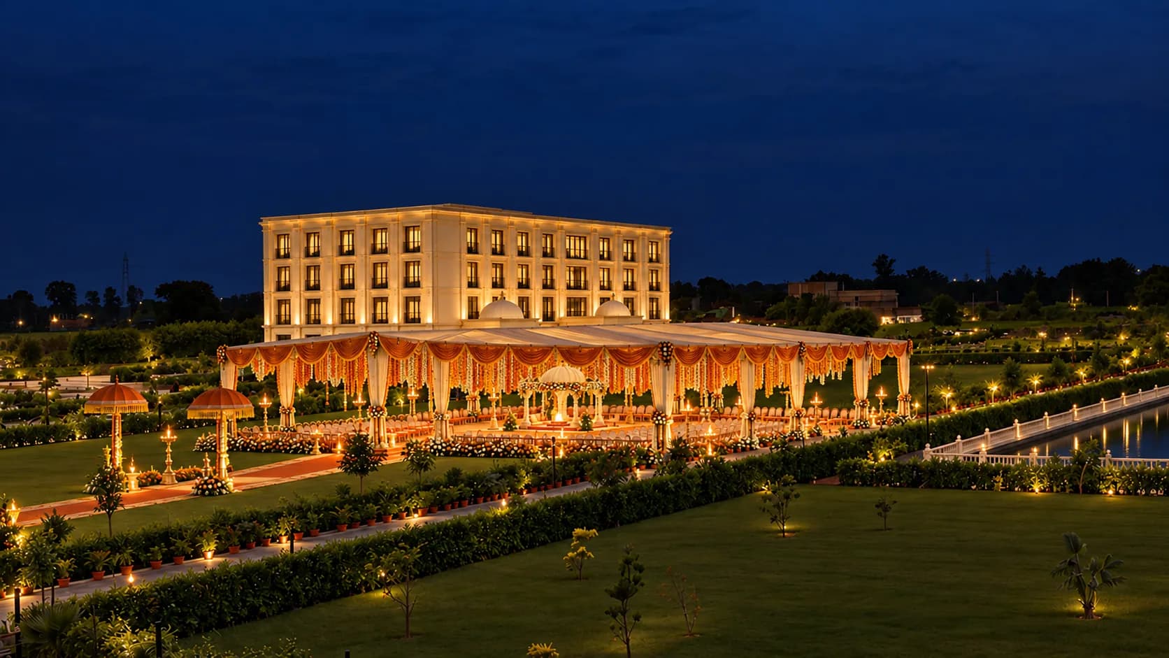 A lawn at night dressed for a wedding: a lit canopy with marigold swags, lamps along the paths and the hotel behind