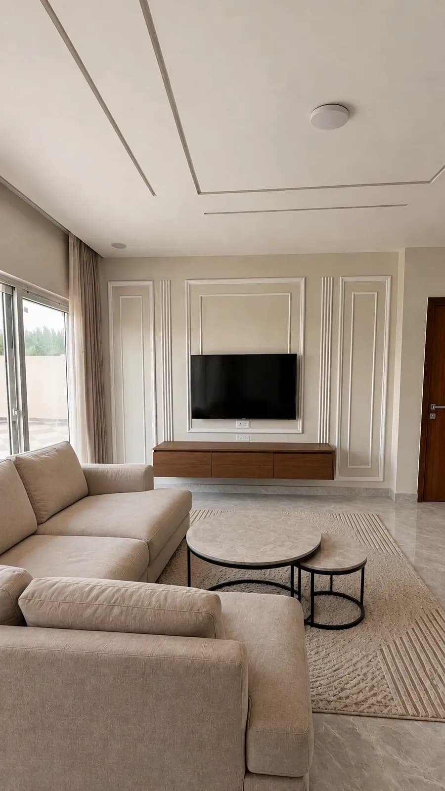 The corner of a villa living room: cream sofas, a round table and a television on a panelled wall